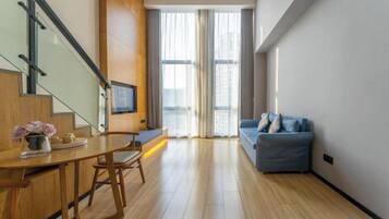 A modern living room with a wooden floor, a blue sofa, a dining table with chairs, and a staircase with a TV on the wall.