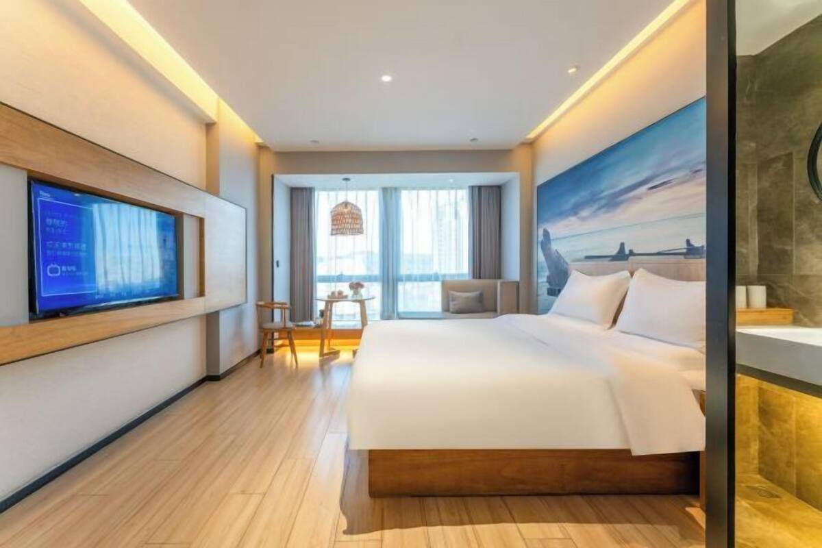 A modern hotel room with a large bed, a television on the wall, a desk with a chair, and a view of the city through the window.