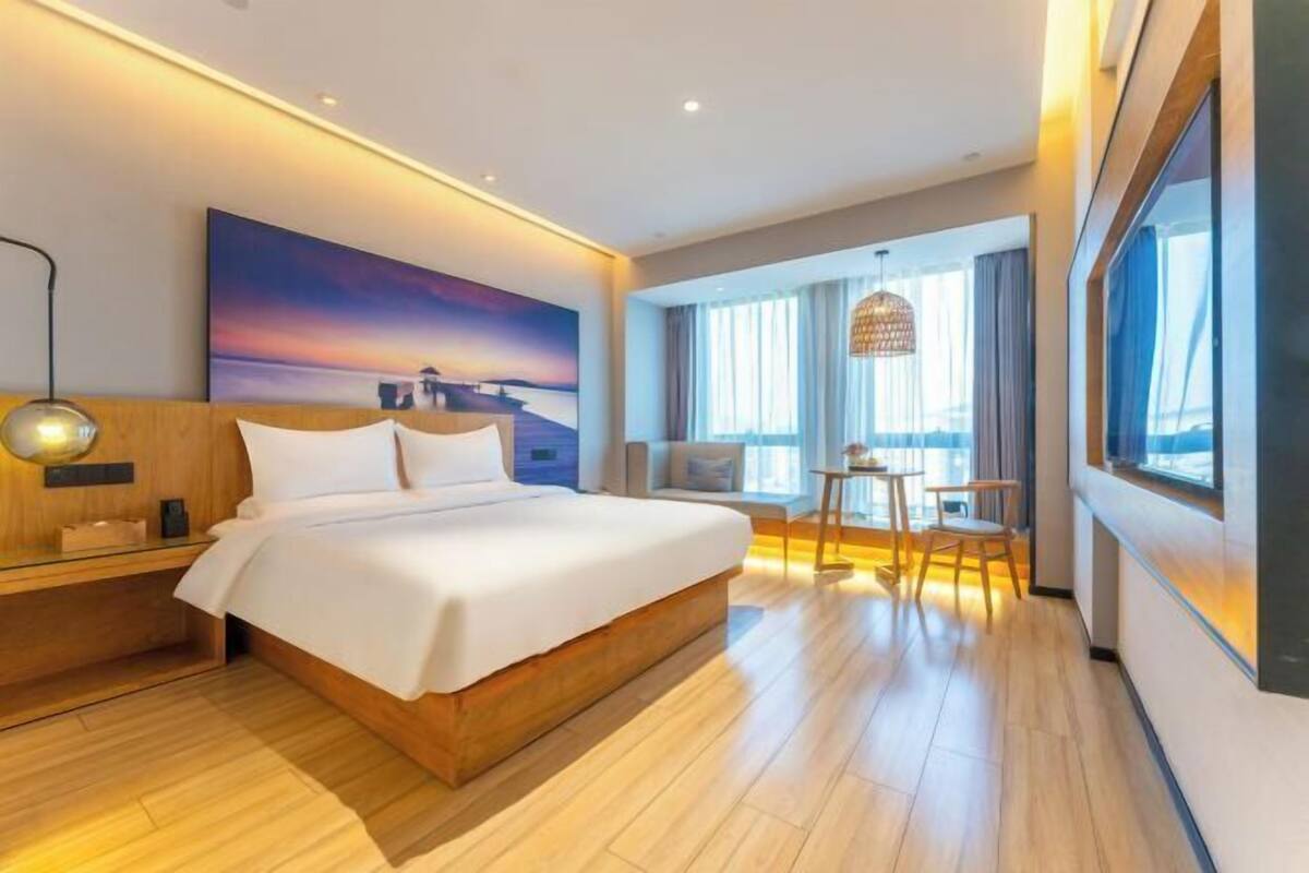 A modern hotel room with a large bed, a bedside table, a desk with a chair, a large window with curtains, and a painting on the wall.