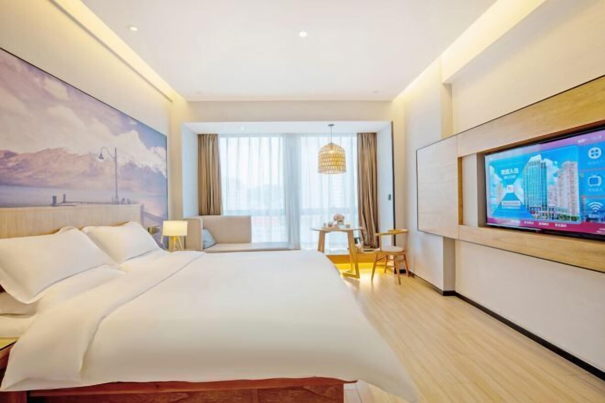 A hotel room with a large bed, a TV mounted on the wall, a desk with a chair, and a view of the cityscape through the window.