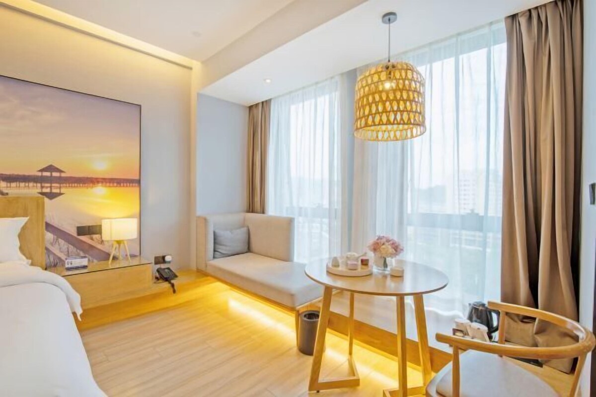 A hotel room with a bed, a sofa, a round table with a vase of flowers, a hanging lamp, and a painting of a sunset.