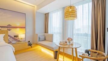 A hotel room with a bed, a sofa, a round table with a vase of flowers, a hanging lamp, and a painting of a sunset.