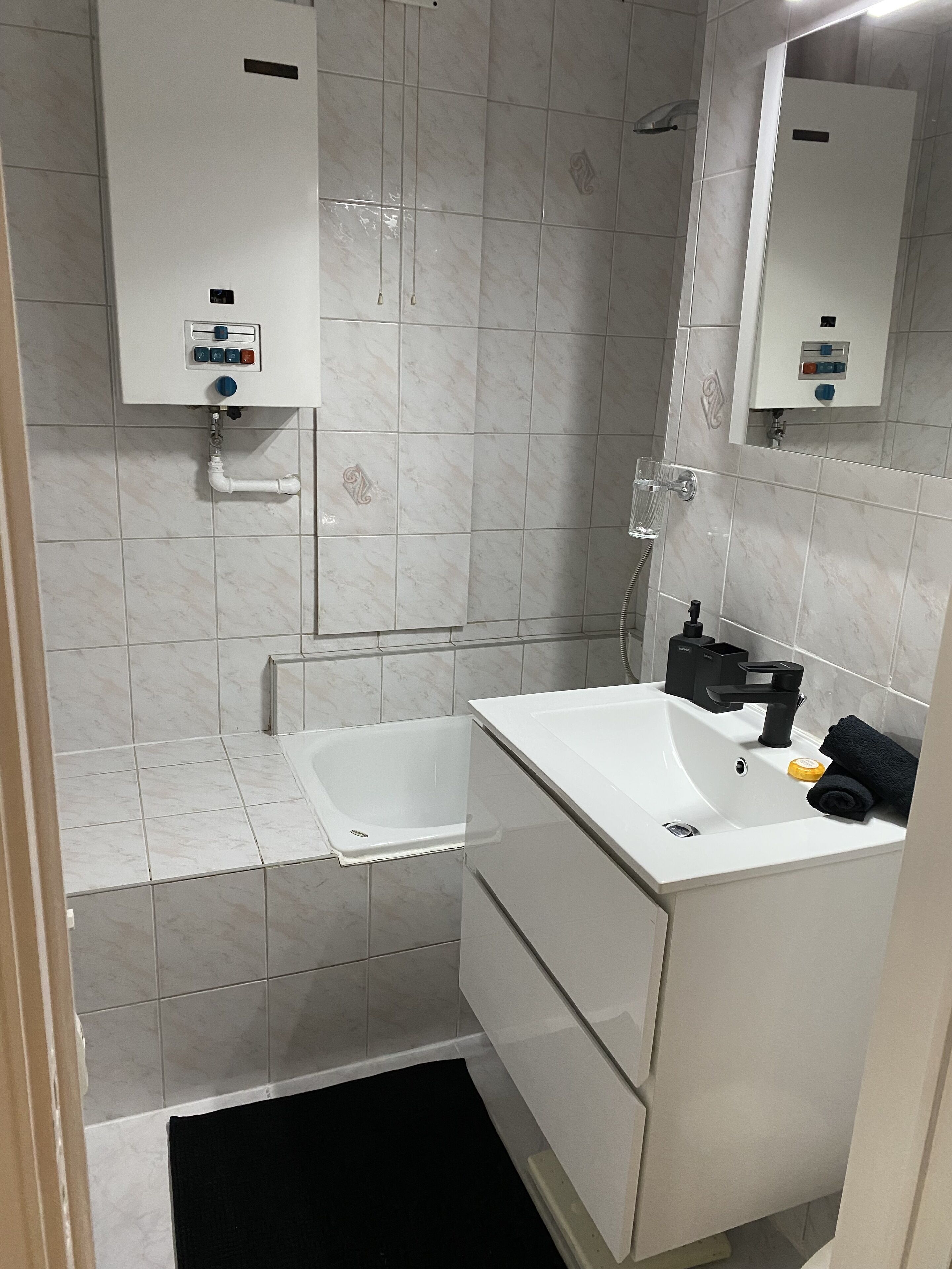 Een badkamer met een wit wastafelmeubel, een bad en een douche. Boven het wastafelmeubel en de douche bevinden zich bedieningspanelen.