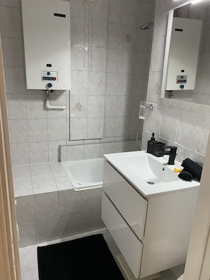 Ein Badezimmer mit einem weißen Waschbecken, einer Badewanne und einer Dusche. Über Waschbecken und Dusche befinden sich Bedienfelder.