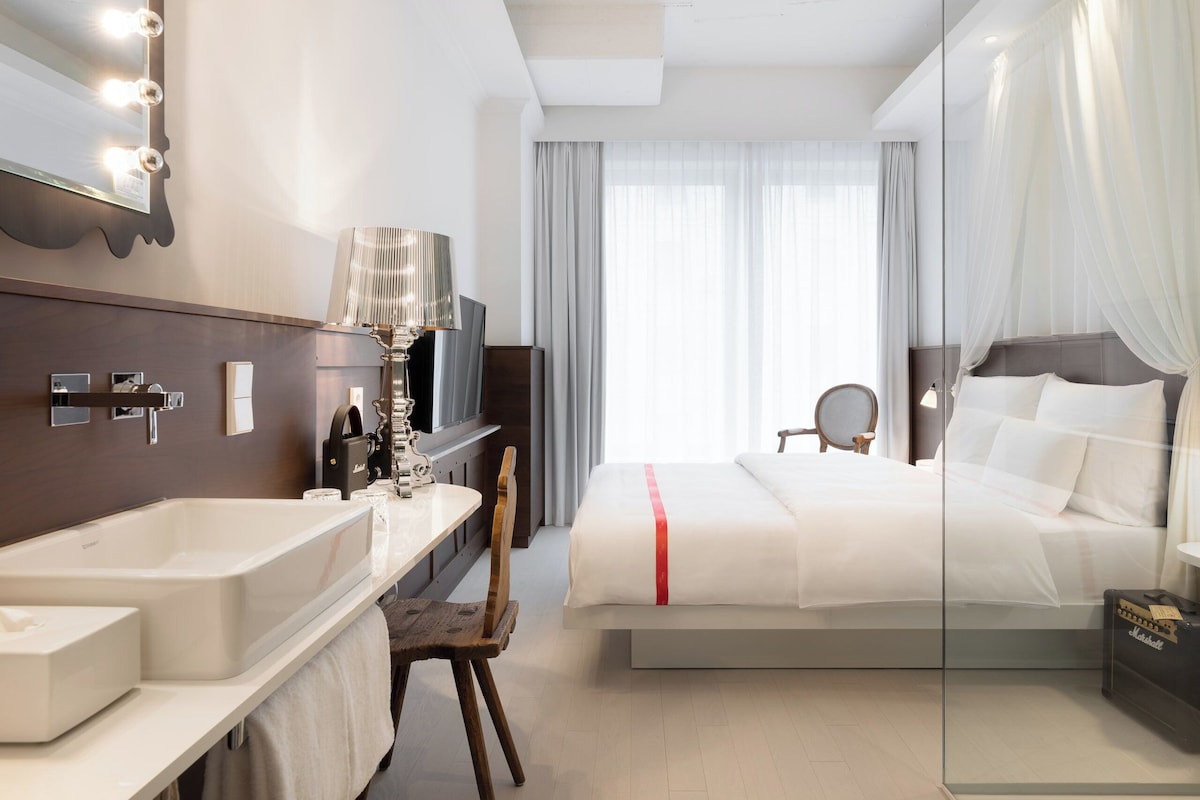 A hotel room with a large bed, a vanity with a mirror and lights, a bathtub, and a television.