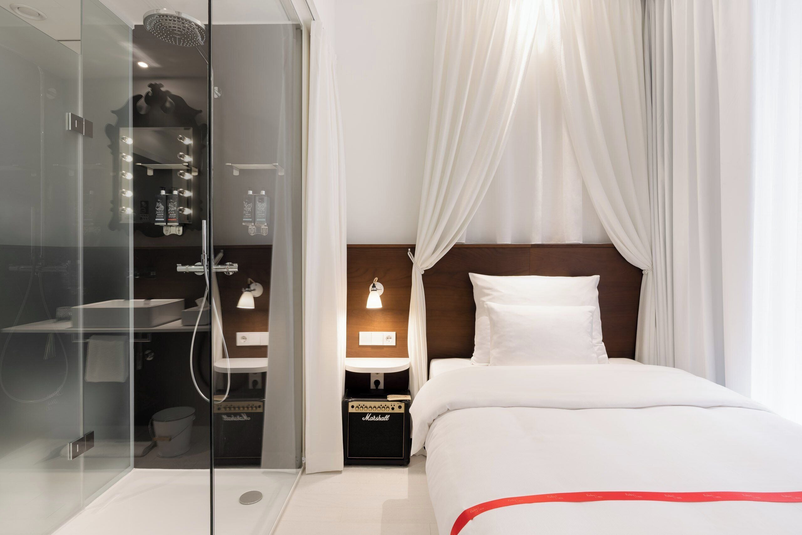 A modern hotel room with a glass-enclosed shower, a vanity with a mirror and lights, a bedside table with a lamp, a bed with white bedding, and a wooden headboard.