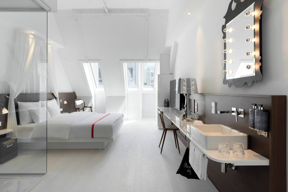 A modern hotel room with a large bed, a vanity with a mirror and lights, a desk, and a glass partition.