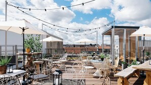 Eine Terrasse im Freien mit Tischen, Stühlen, Lichterketten und Sonnenschirmen.