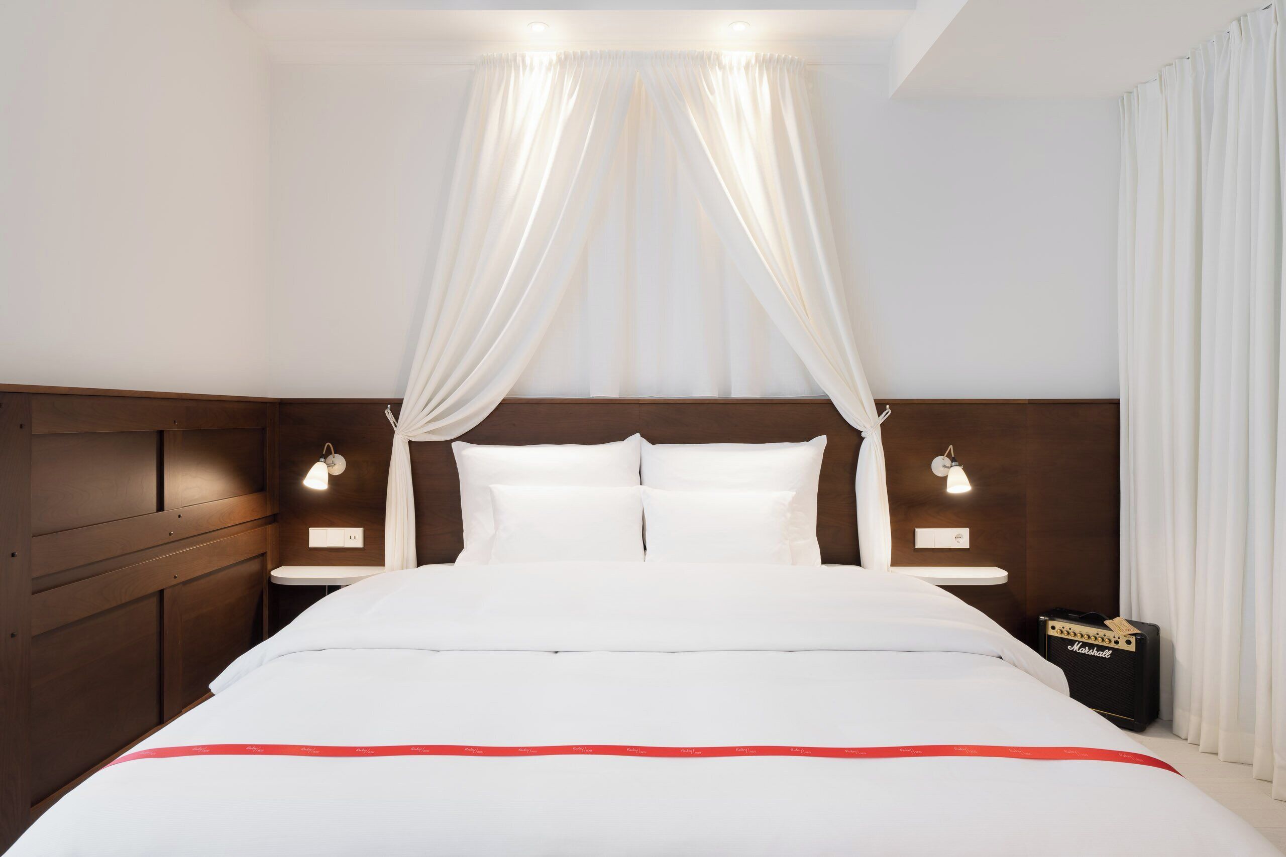 Une chambre d’hôtel avec un grand lit, deux lampes de chevet, un amplificateur Marshall et un baldaquin au-dessus du lit.