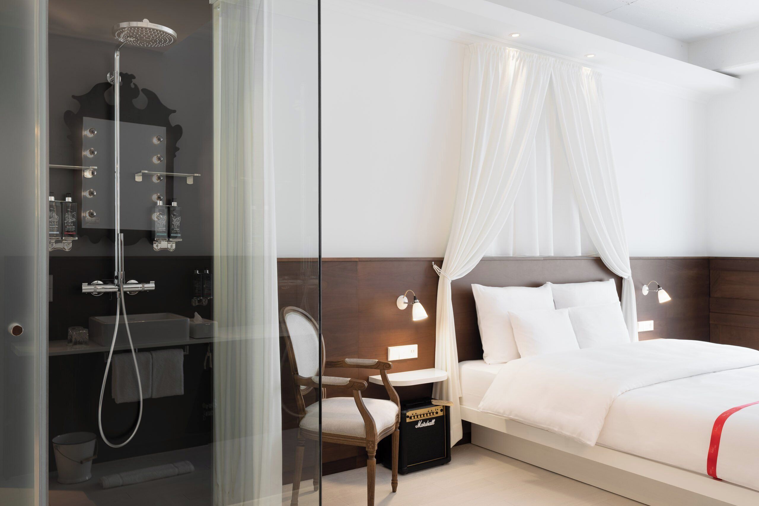 Une chambre d’hôtel dotée d’une douche en verre, d’un lit avec une literie blanche, d’une table de chevet, d’une chaise et d’une petite table de nuit avec une lampe.