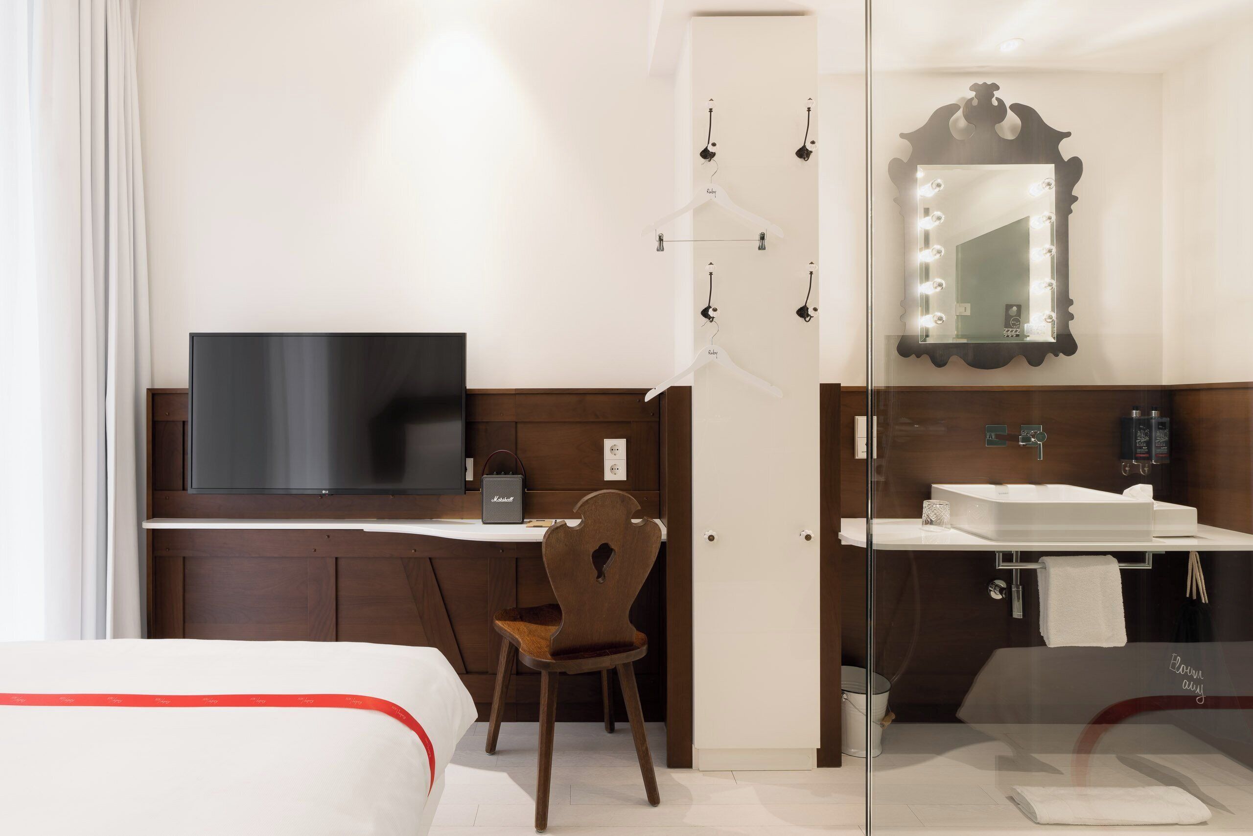 A hotel room with a bed, a desk with a chair, a television, a mirror with lights, and a bathroom with a sink and mirror.
