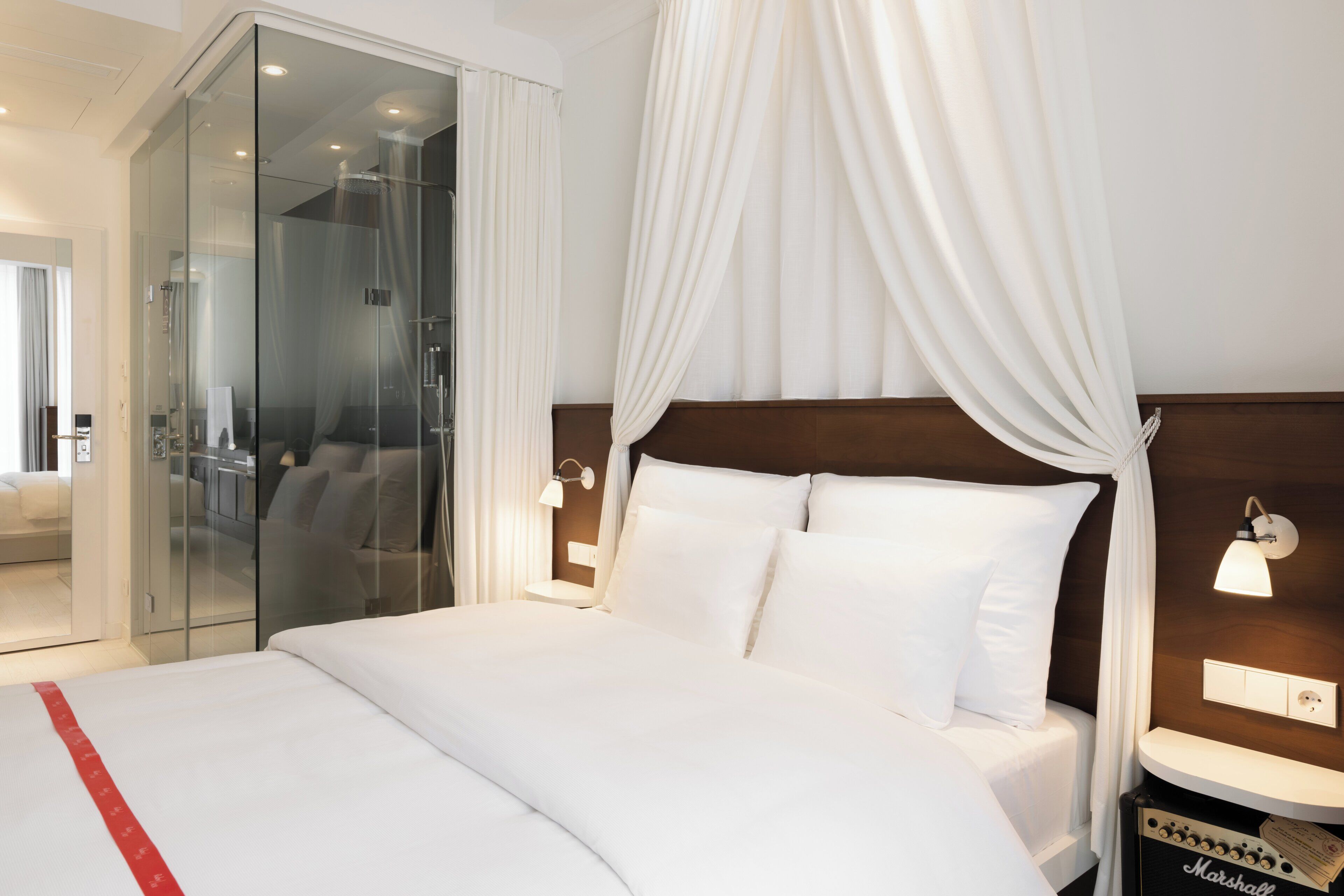 A hotel room with a large bed, a glass shower enclosure, a bedside table with a lamp, and a Marshall amplifier on the nightstand.