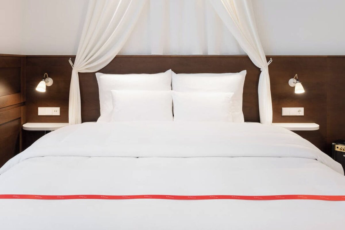 A hotel room with a bed, white curtains, bedside lamps, and a wooden headboard.