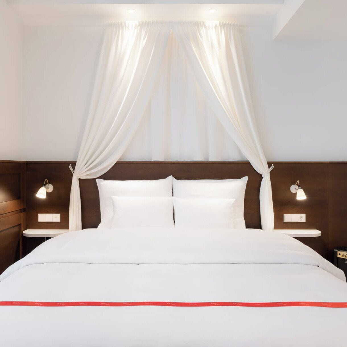 A hotel room with a bed, white curtains, bedside lamps, and a wooden headboard.