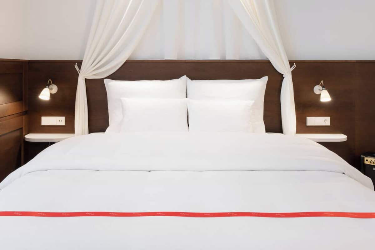 A hotel room with a bed, white curtains, bedside lamps, and a wooden headboard.