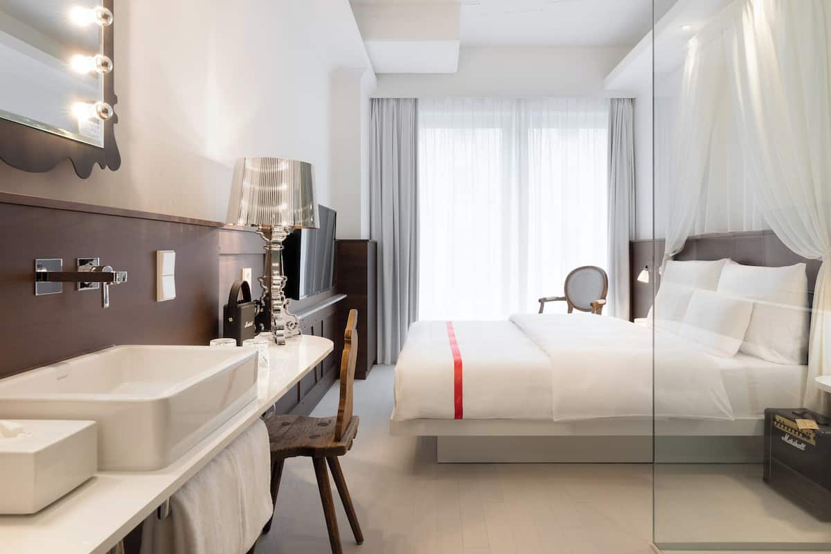 A hotel room with a large bed, a vanity with a mirror and lights, a bathtub, and a television.