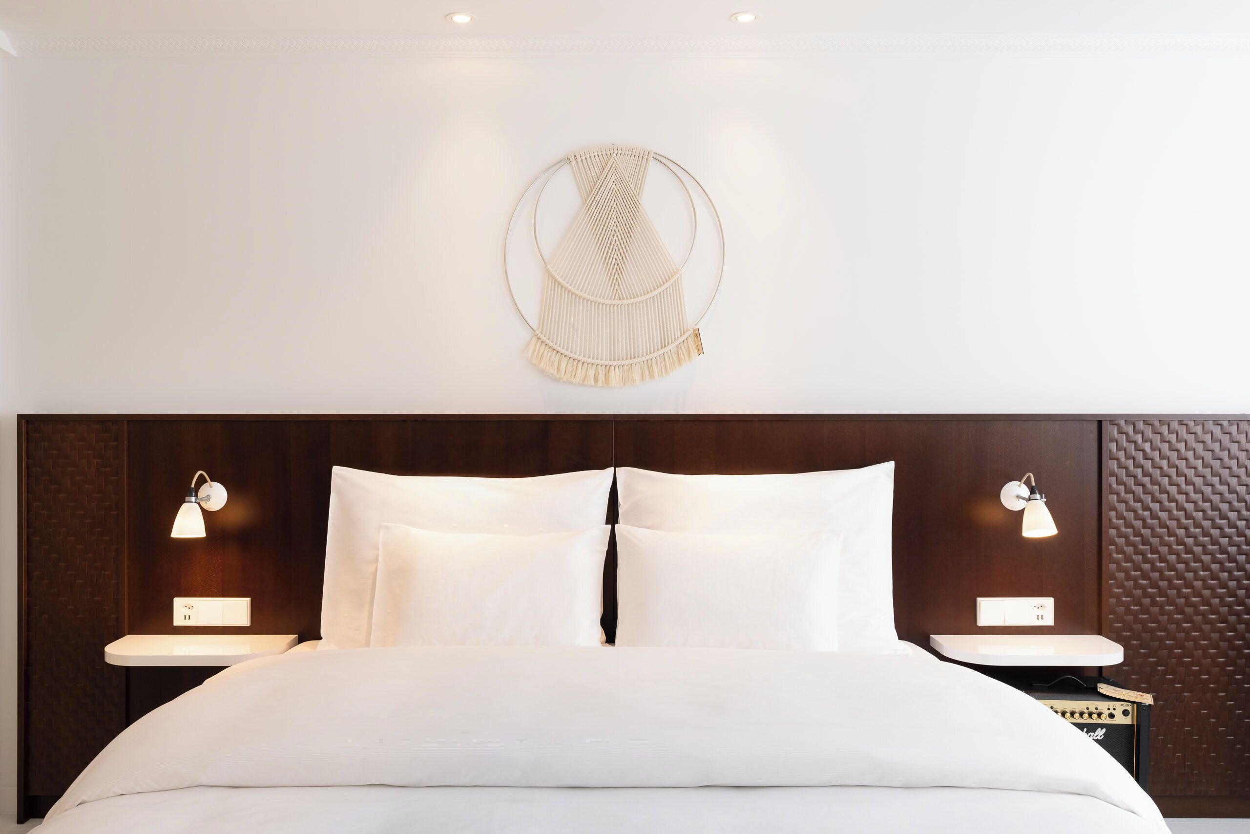Habitación de hotel con cama, dos mesitas de noche con lámparas, un tapiz de macramé en la pared y un panel mural texturizado.