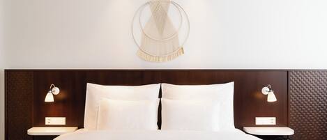Habitación de hotel con cama, dos mesitas de noche con lámparas, un tapiz de macramé en la pared y un panel mural texturizado.