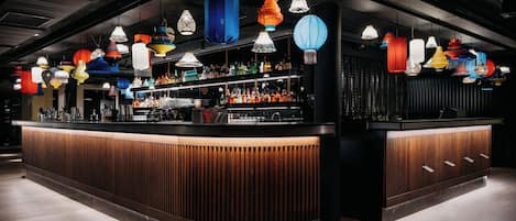 Un bar doté d’un comptoir en bois aux lignes courbes et agrémenté de différents luminaires suspendus aux couleurs vives.