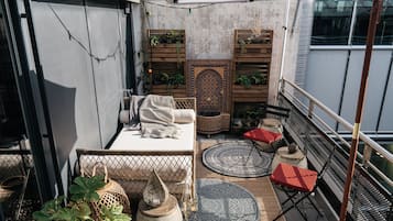 Un pequeño balcón con un sofá, un otomano estampado, una silla y un mueble decorativo con plantas.