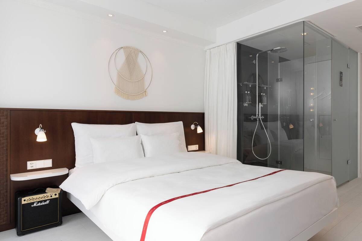 Une chambre d’hôtel moderne avec un grand lit, une douche en verre, un luminaire fixé au mur et un petit haut-parleur.