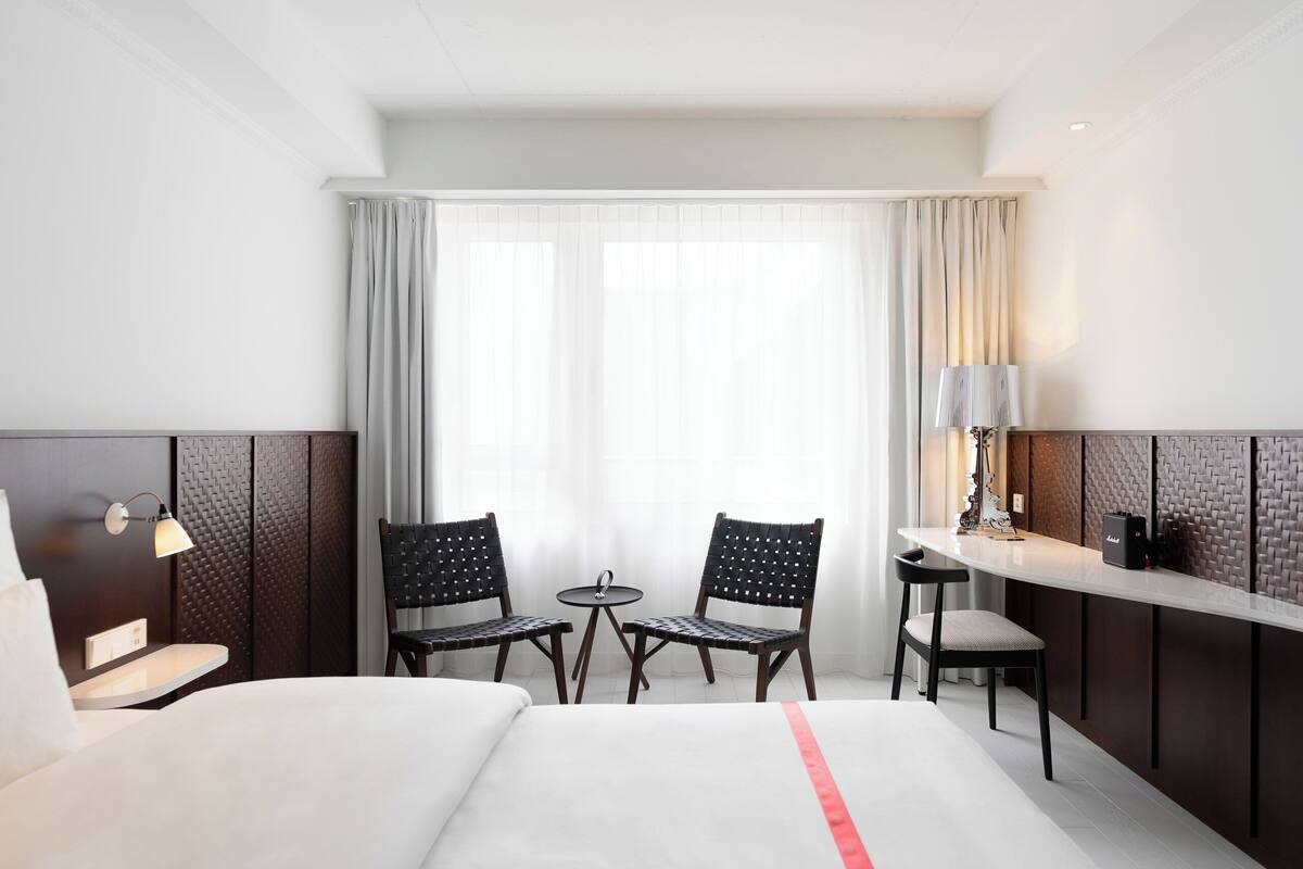 Une chambre d’hôtel avec un lit, deux chaises, une petite table, une lampe et une vue sur l’extérieur par une fenêtre.