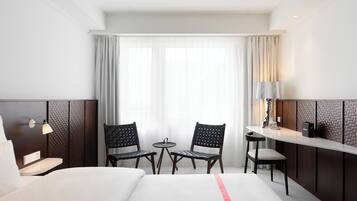 A hotel room with a bed, two chairs, a small table, a lamp, and a view of the outside through a window.