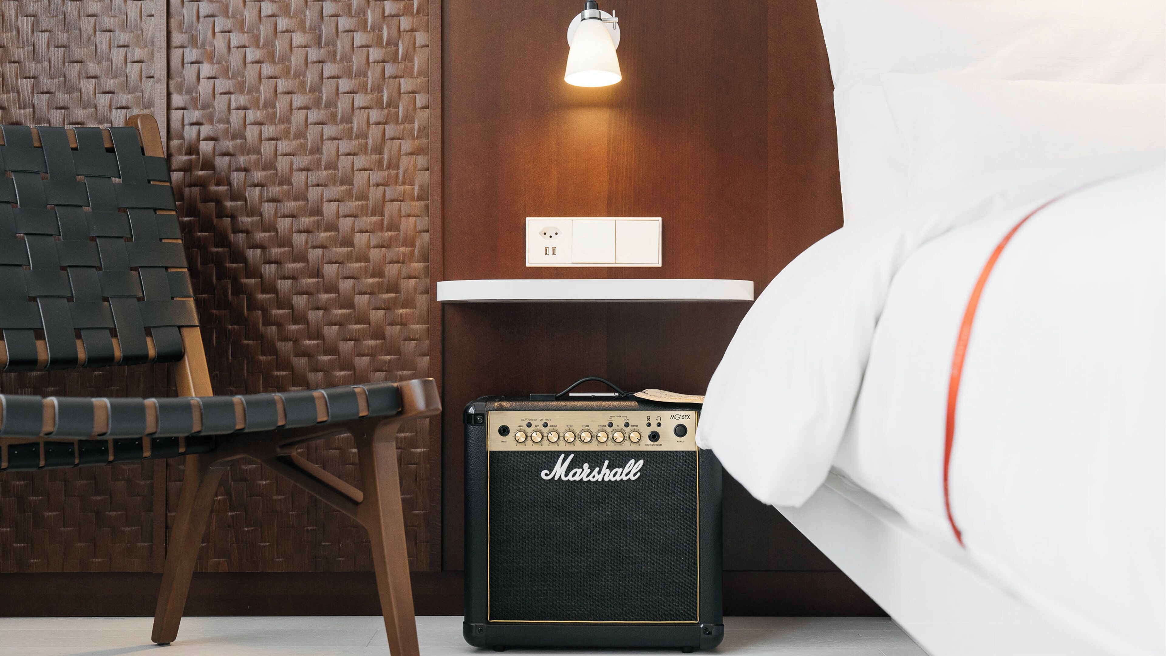 Une pièce avec un mur recouvert d’un revêtement tissé, une chaise, une petite table avec une lampe et un amplificateur Marshall.
