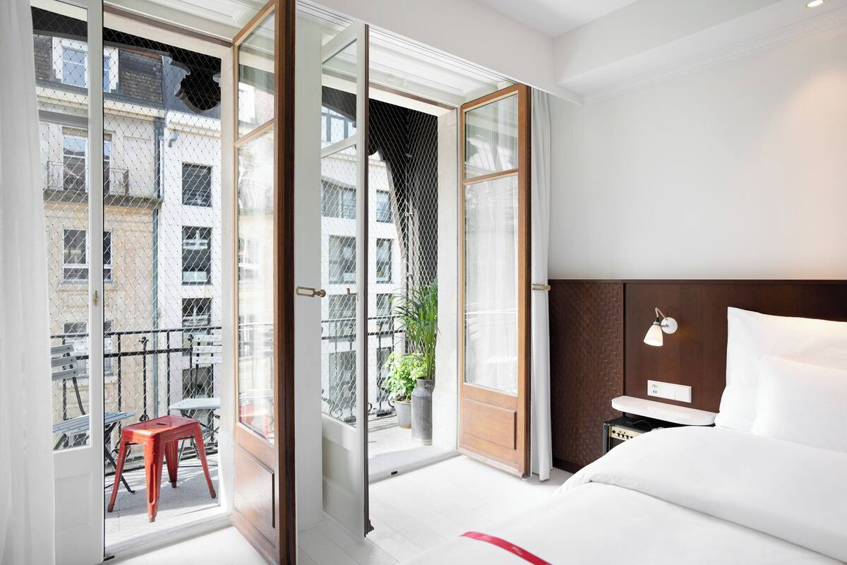 Une chambre d’hôtel comprenant un lit, une table de chevet, une lampe, une porte-fenêtre coulissante donnant sur un balcon extérieur et une vue sur les bâtiments environnants.
