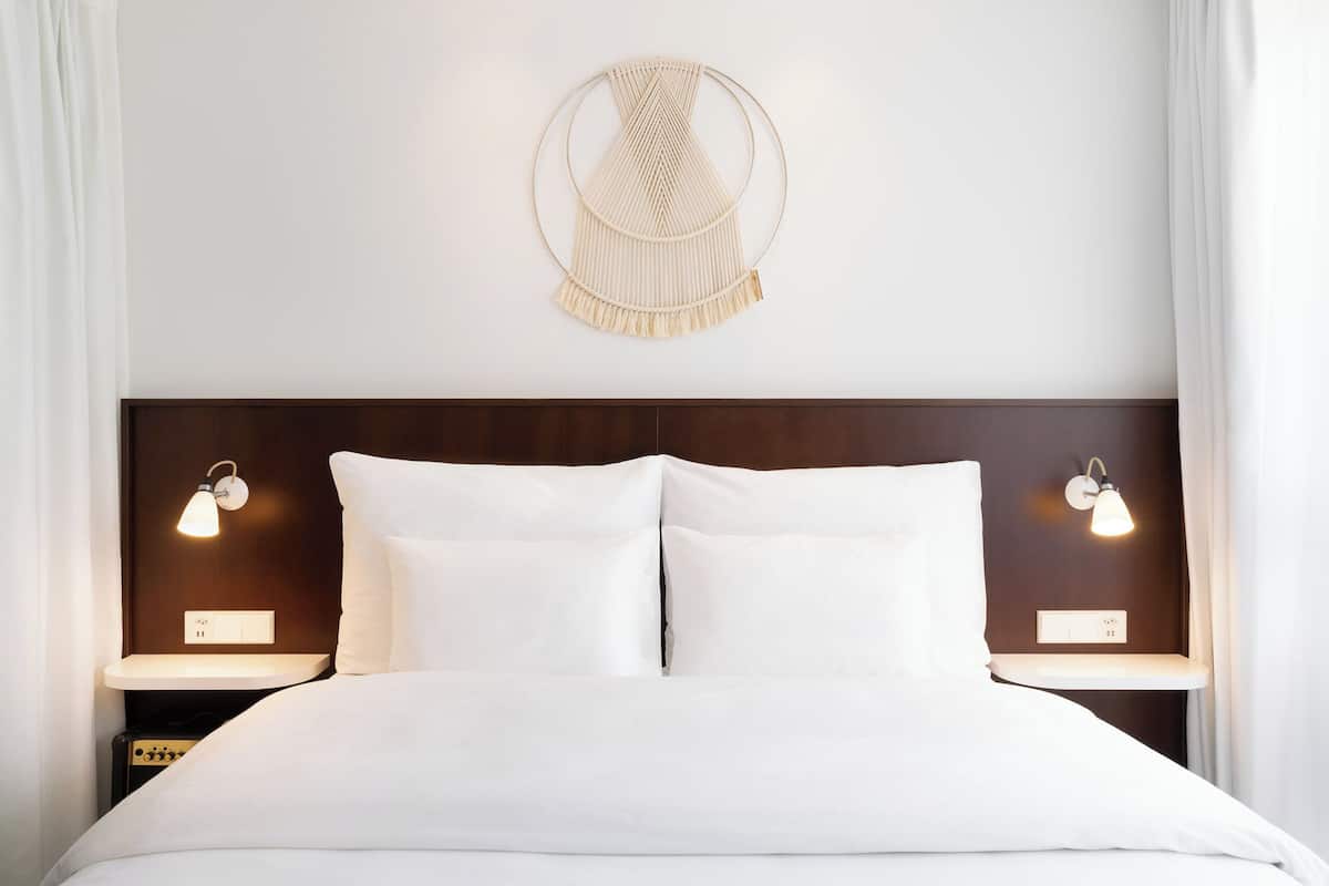 Un lit avec une literie blanche, deux lampes de chevet, une décoration murale en macramé et une tête de lit en bois foncé.