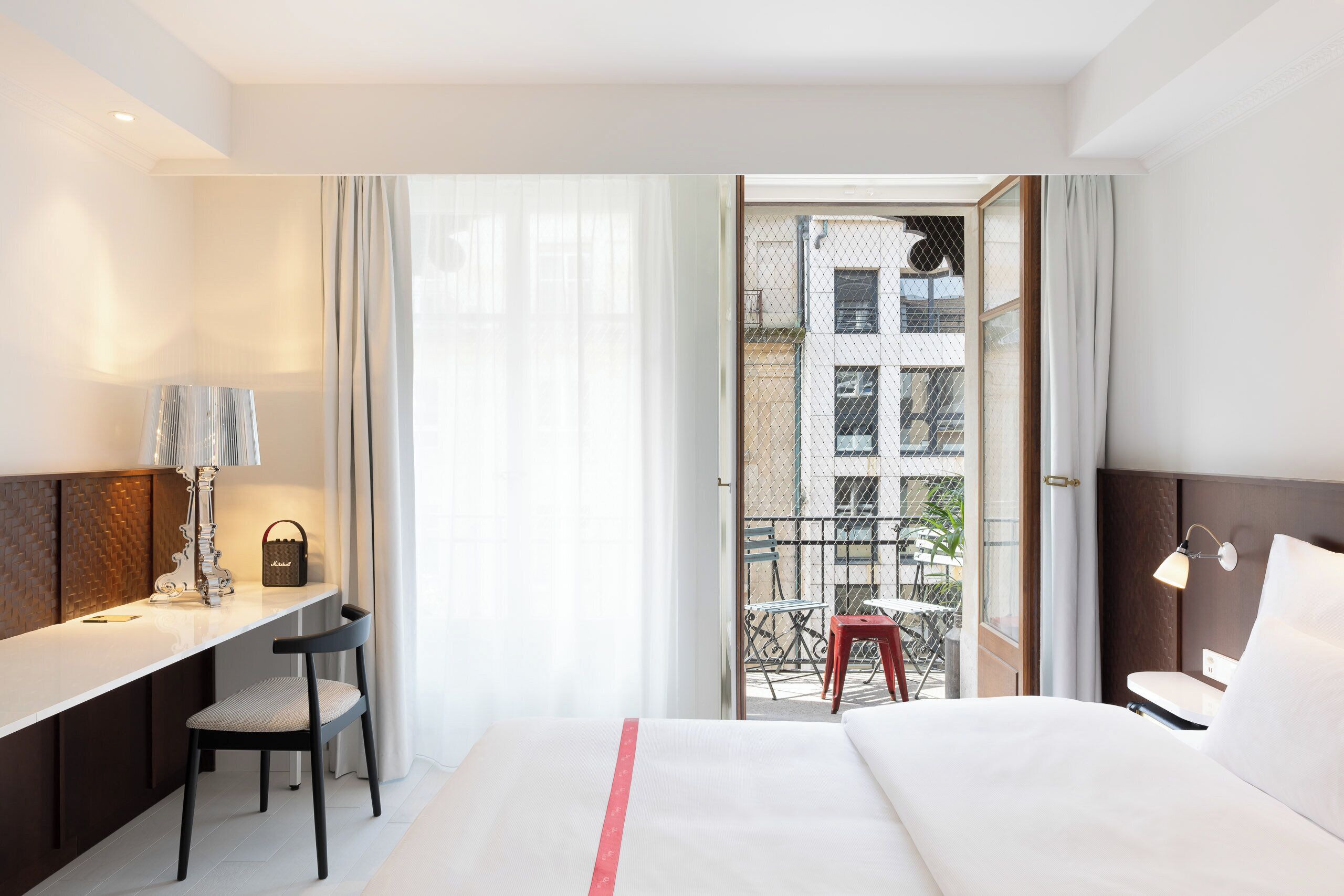 A hotel room with a bed, a desk, a chair, a lamp, and a view of the outside.