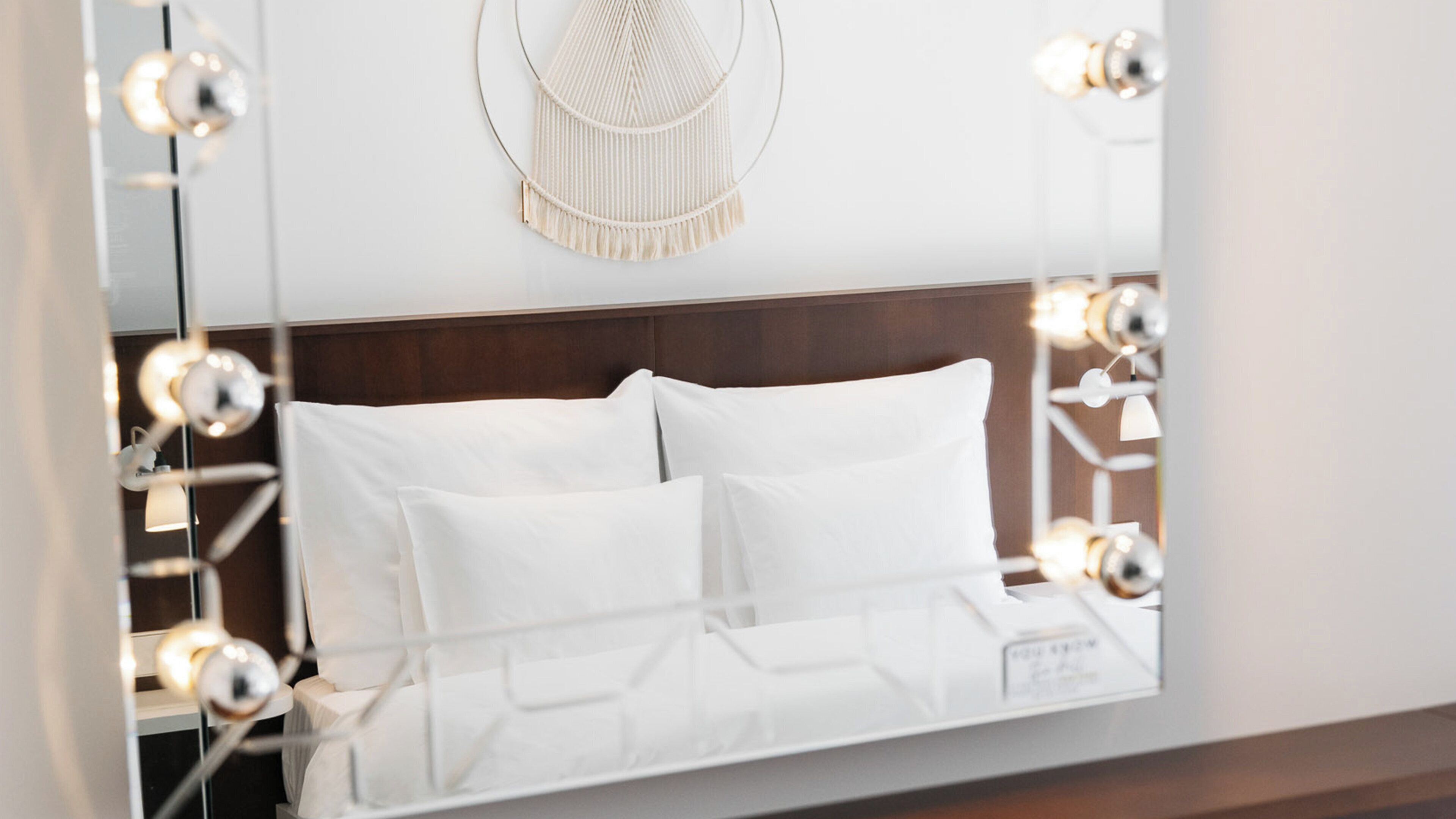 A bed with white linens, a wooden headboard, and a macrame wall hanging above.