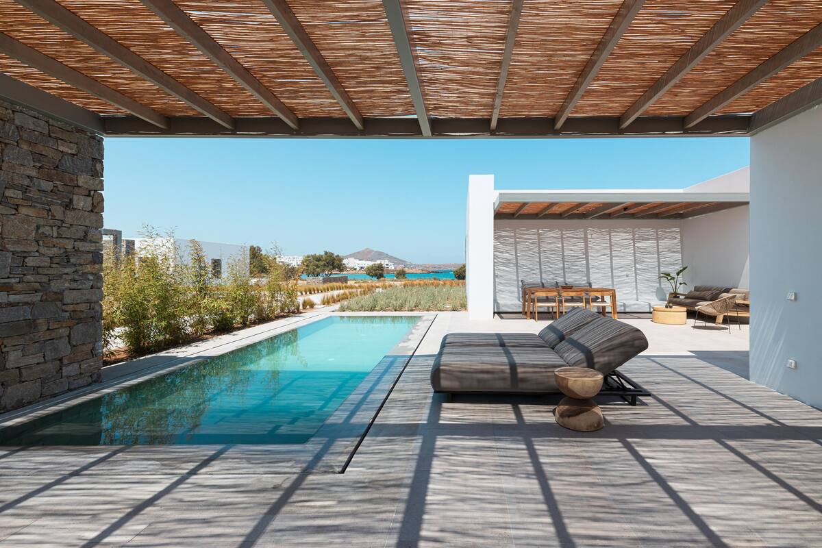 Un área de piscina moderna al aire libre con un muro de piedra, una pérgola de madera y un espacio de estar con cojines y una mesa.