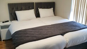 A double bed with a dark headboard and footboard, white bedding, and a dark quilt. There is a bedside table with a remote control on it. The room has wooden flooring and a window with blinds. - City Centre, Detached 10 Person Accommodation (Liverpool)