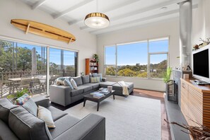 A spacious living area with a large sofa set, a coffee table, and a television. There is a bookshelf and a surfboard hanging on the wall. - Tree Top House (Fairhaven)