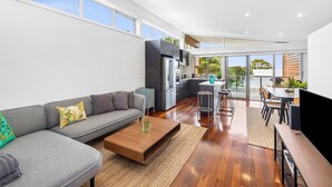 A modern living room with a grey sofa, wooden coffee table, and a kitchen area with dark cabinets and stainless steel appliances.