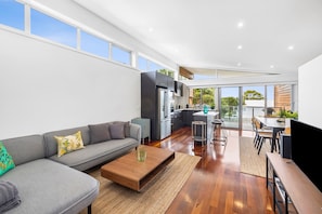A modern living room with a grey sofa, wooden coffee table, and a kitchen area with dark cabinets and stainless steel appliances.