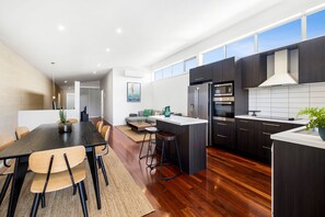 A modern kitchen with dark cabinetry, a central island, and a dining area with a wooden table and chairs. - Beach Baby (Aireys Inlet)