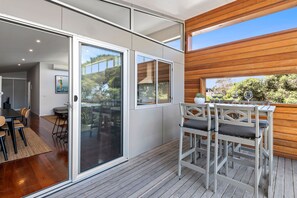 A modern outdoor dining area with a wooden deck, a glass door leading to an indoor space, and a view of the outdoors. - Beach Baby (Aireys Inlet)