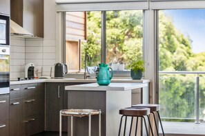 A modern kitchen with a central island, stainless steel appliances, and a view of greenery through large windows. - Beach Baby (Aireys Inlet)