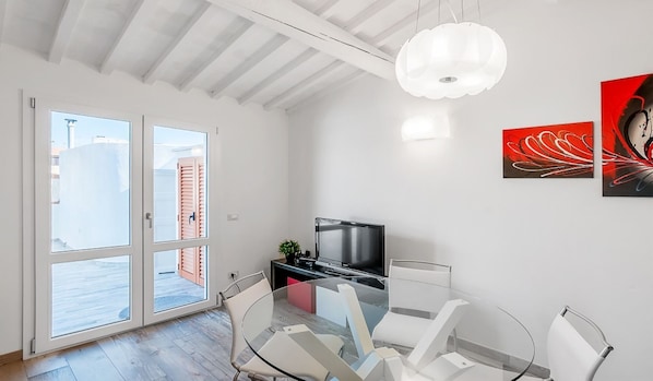 Un salon moderne avec une table à manger en verre, des chaises blanches, un téléviseur à écran plat et un luminaire suspendu au plafond.