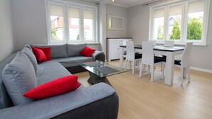 Una sala moderna con un sofá gris, cojines rojos, una mesa de centro de vidrio y un comedor con sillas blancas y una mesa.
