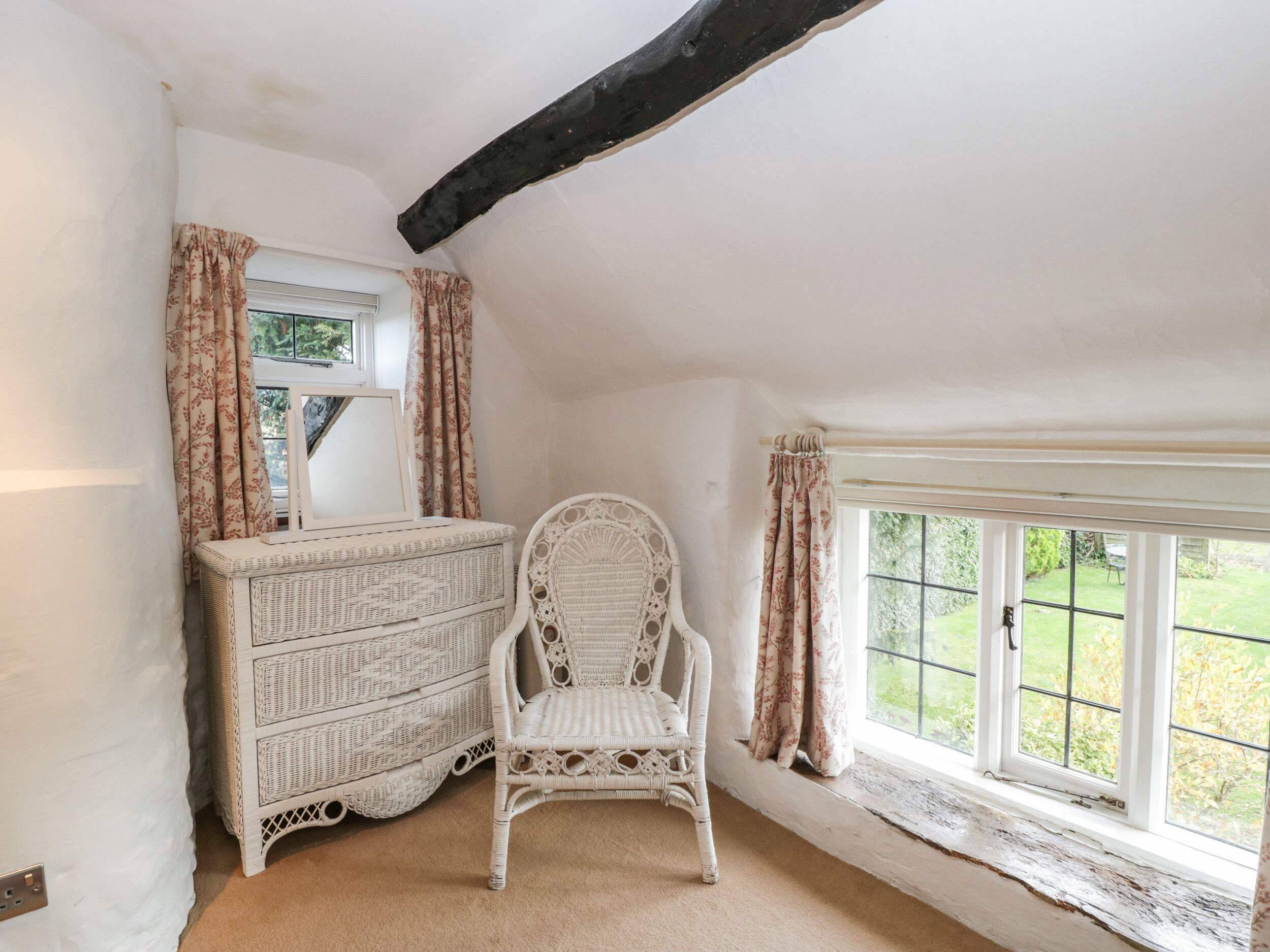 A room with a white wicker chest of drawers, a white wicker chair, and a large window with curtains.