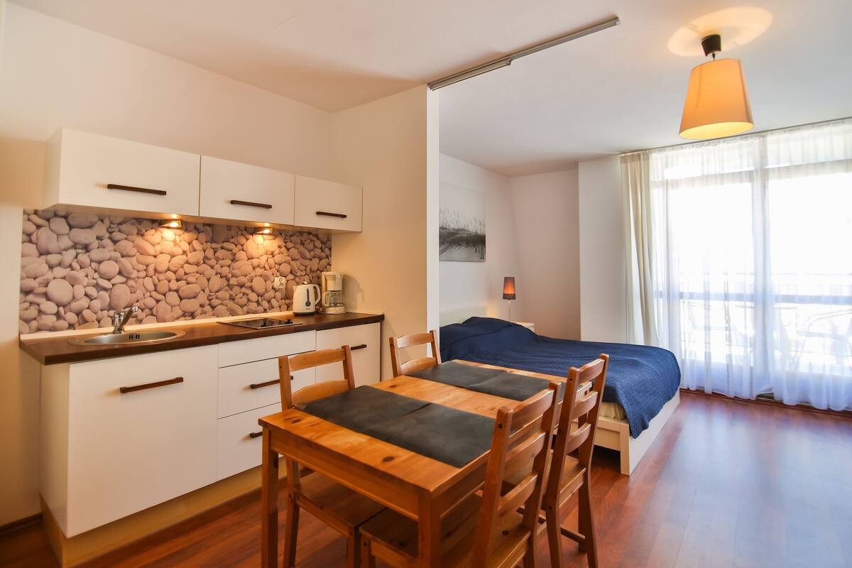 Una habitación compacta con kitchenette, una mesa de comedor con sillas, una cama con cubrecama azul y un ventanal con cortinas translúcidas.