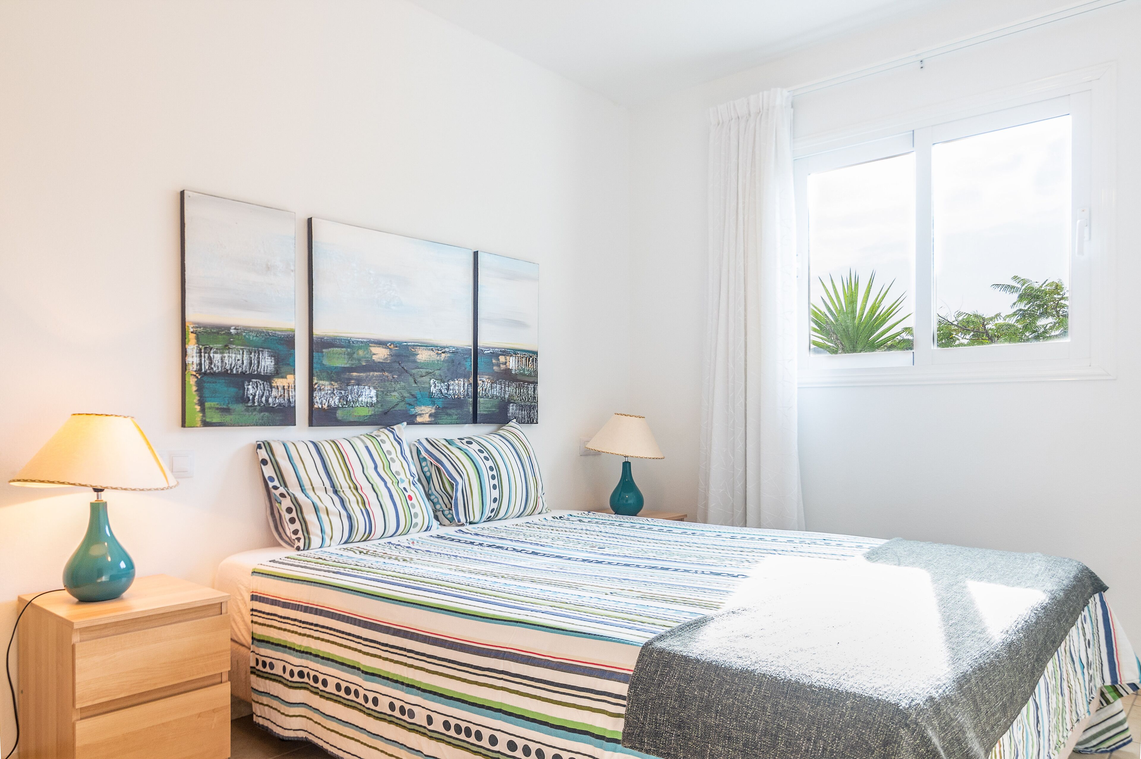 Un dormitorio con cama, dos lámparas de noche, una mesita de noche y ventana con vista a follaje.