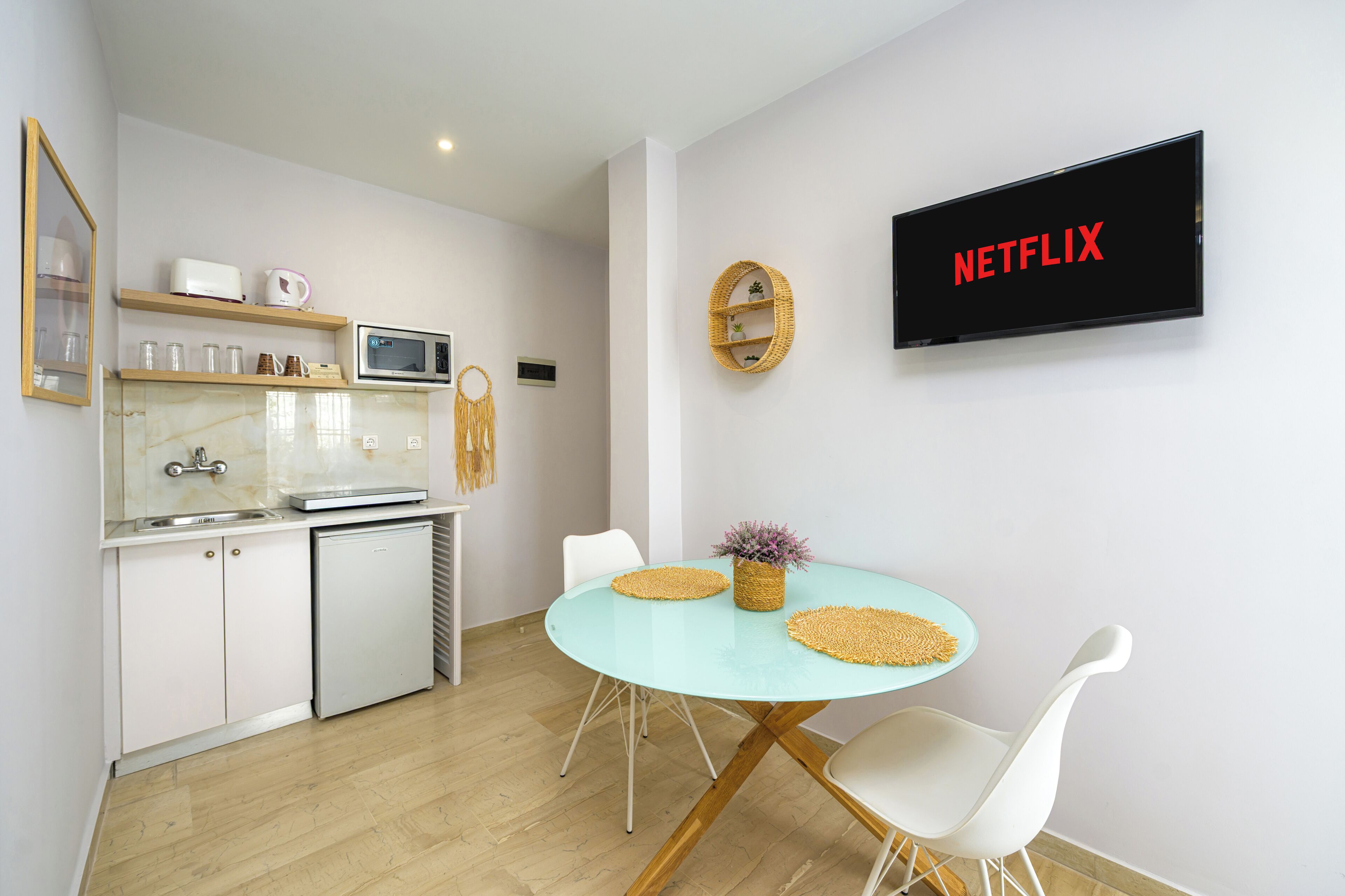 A modern kitchen with white cabinets, a microwave, and a coffee maker. There is a dining area with a round table and white chairs. A TV mounted on the wall displays "NETFLIX".