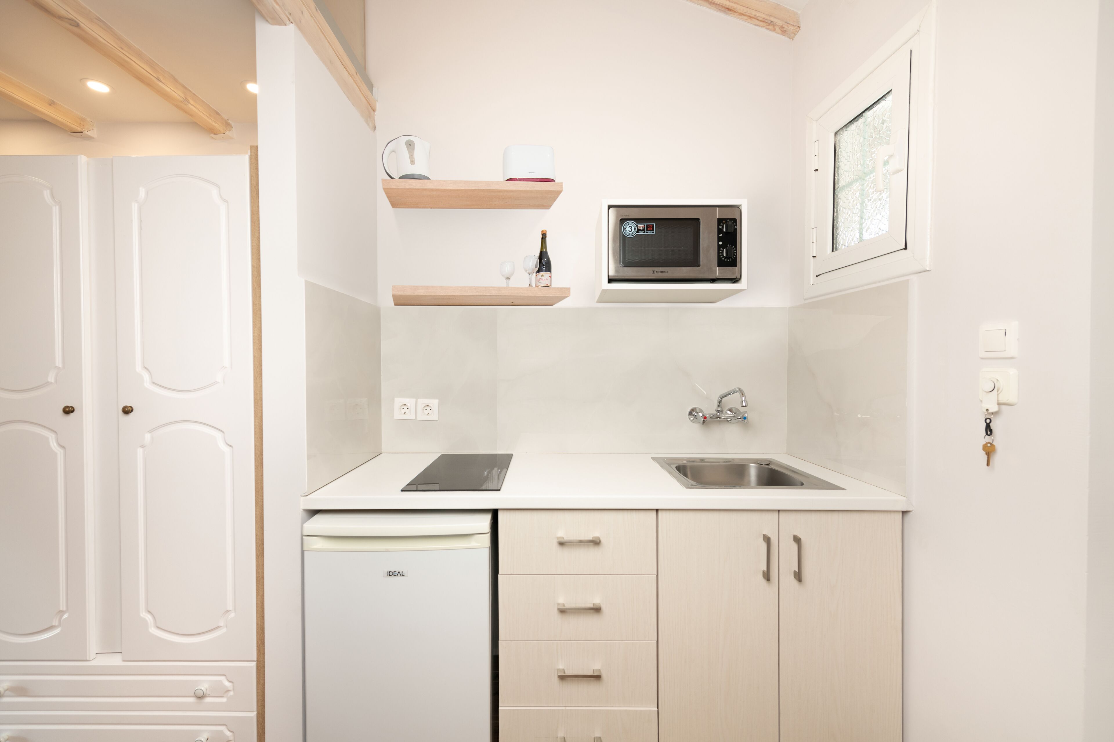A compact kitchen with a dishwasher, microwave, and a sink. There are wooden shelves above the microwave and a door with a handle on the left.