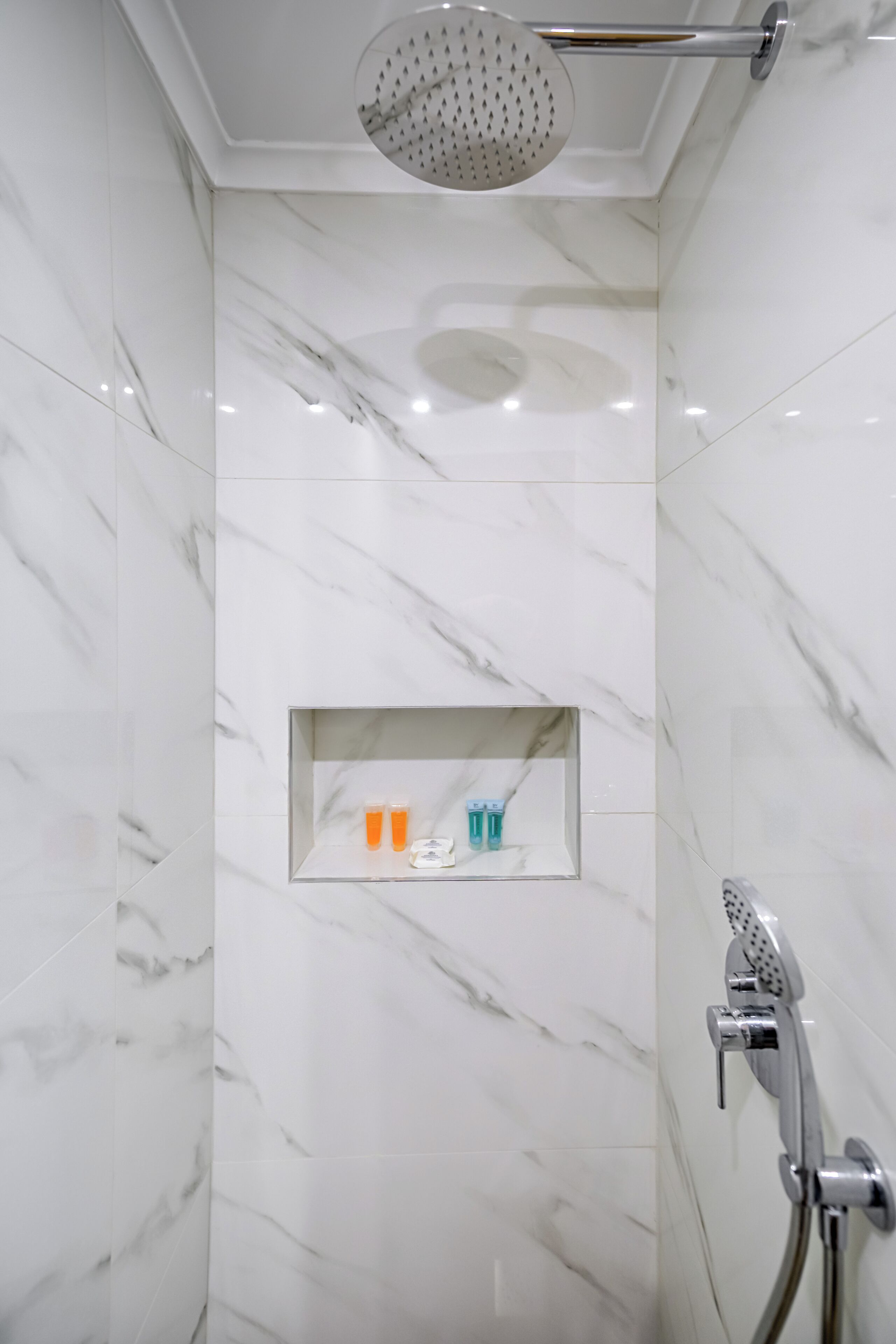 A bathroom with white marble walls and a built-in shelf with toiletries.