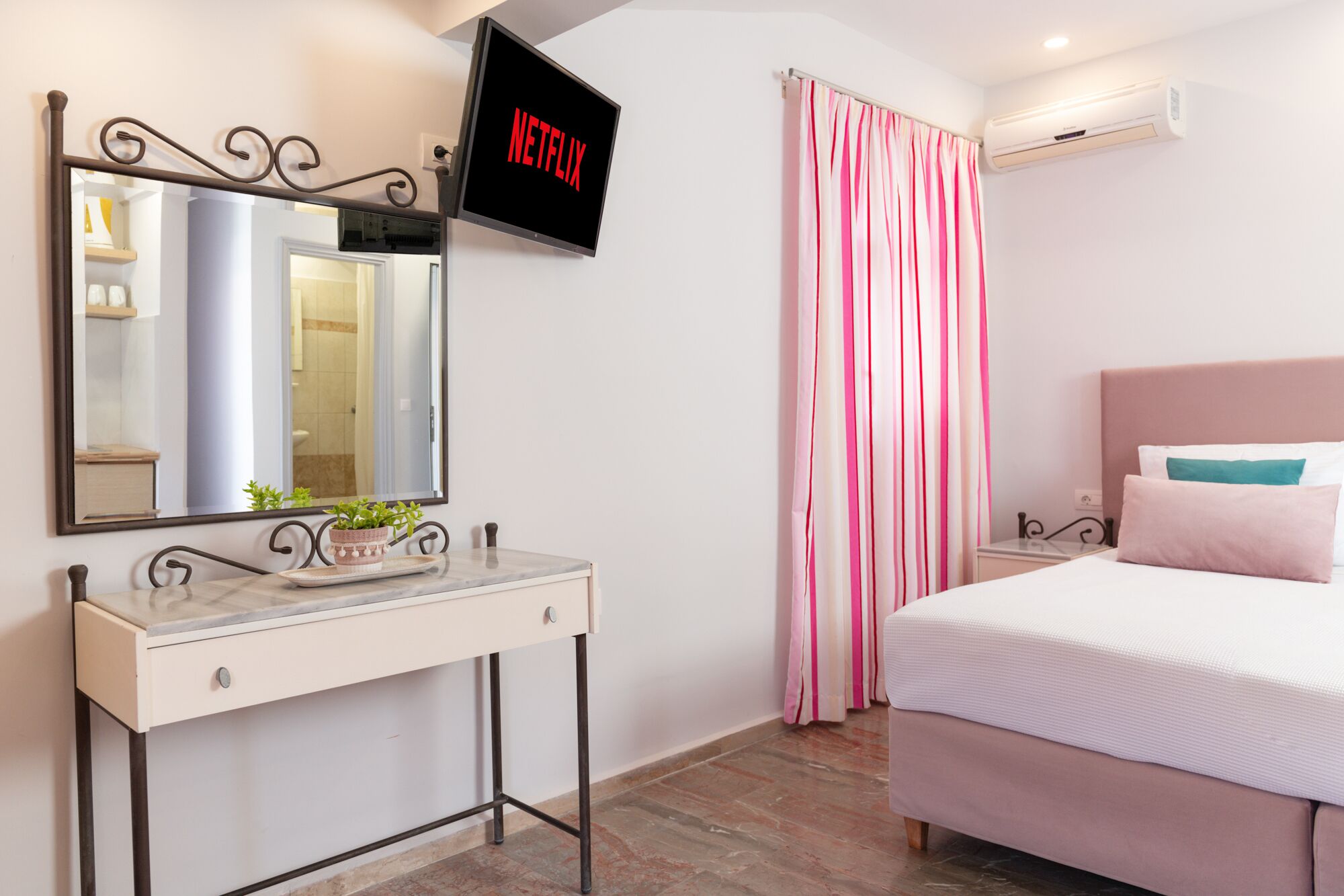 Une chambre à coucher comprenant un lit, une table de chevet avec un miroir, un téléviseur fixé au mur et une fenêtre avec des rideaux.