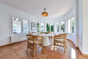 A bright dining room with a round table, wicker chairs, and large windows offering a view of greenery.